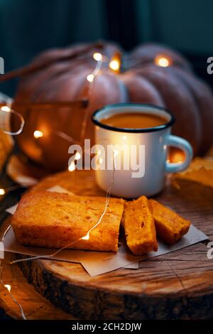 Bella composizione di festa con luci per Natale e carte di ringraziamento. Una tazza di stagno con una bevanda e una torta appena sfornata su un supporto di legno. Una zucca grande sullo sfondo. Foto Stock