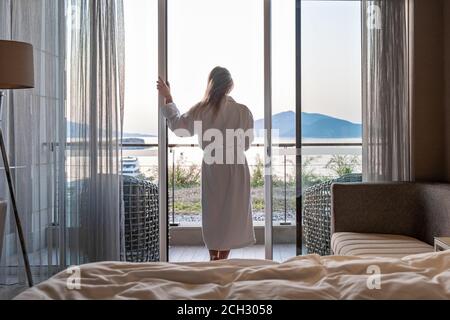Bella donna dal retro in accappatoio in piedi sulla terrazza di Camera d'hotel con vista mare alba mattina Foto Stock