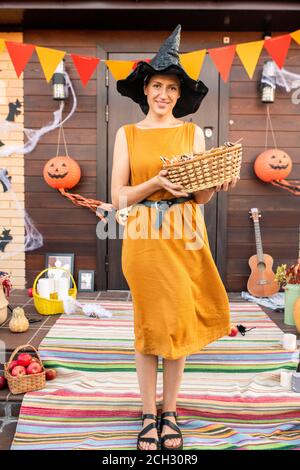 Giovane donna sorridente in elegante abito e tenuta cappello nero cestino con dolci Foto Stock