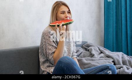 Giovane donna attraente morde un pezzo di anguria. Donna a casa in un interno accogliente Foto Stock