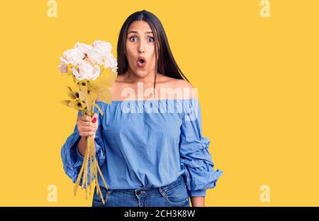 Giovane bella donna brunette che tiene bouquet spaventato e stupito con bocca aperta per sorpresa, viso incredulo Foto Stock