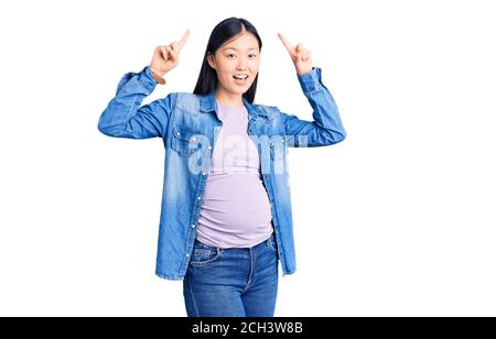 Giovane bella donna cinese incinta aspettandosi bambino sorridente stupito e sorpreso e puntando in su con le dita e le braccia sollevate. Foto Stock