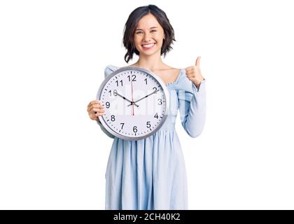 Giovane bella ragazza tenendo grande orologio sorridente felice e positivo, pollice su facendo eccellente e segno di approvazione Foto Stock