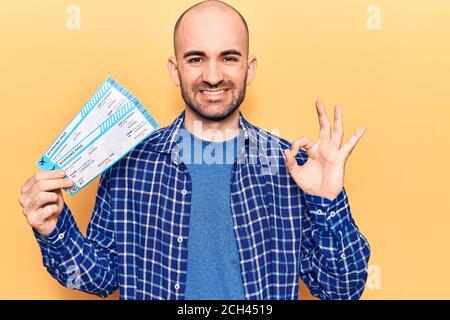 Giovane bel calvo uomo che tiene la carta d'imbarco della compagnia aerea facendo ok segno con le dita, sorridente amichevole gesturing simbolo eccellente Foto Stock