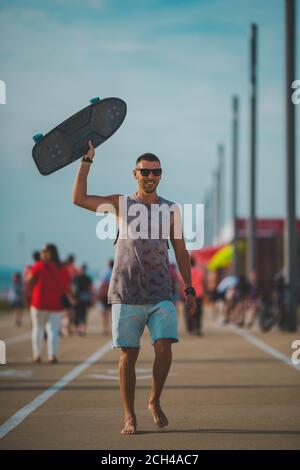 Felice uomo sportivo spensierato godendo la vita e camminando con la longboard in mano al tramonto. Concetto di attività estive Foto Stock