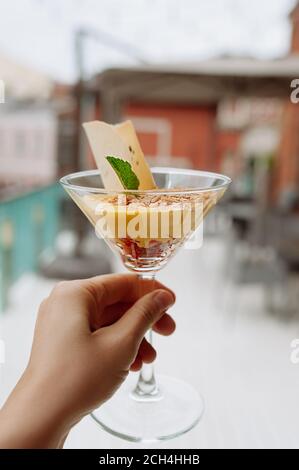 tenere a mano un elegante bicchiere con dolce mousse giallo e biscotti contro la terrazza esterna del ristorante, fuoco selettivo, profondità di campo, verticale Foto Stock