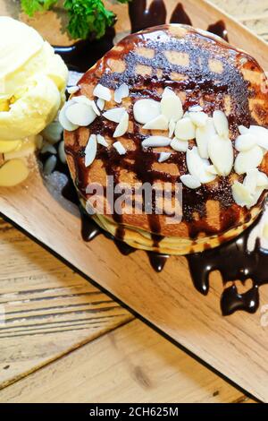 Vista dall'alto dei pancake alla banana con noci di macadamia e sciroppo di cioccolato sulla pila di frittelle e gelato alla vaniglia su un piatto di legno su un tavolo di legno. Foto Stock