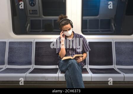 Un giovane adulto isolato e mascherato che ascolta musica e lettura di un libro di letteratura mentre si viaggia in metropolitana Foto Stock