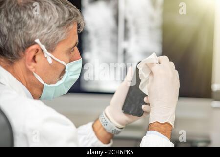 Uomo che pulisce il suo telefono indossando maschera e guanti in ospedale. Foto Stock