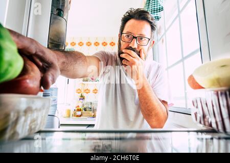 Uomo caucasico adulto che guarda all'interno del frigorifero pensando e scegliendo cosa mangiare per pranzo o cena a casa - concetto di persone e cibo per la dieta e wei Foto Stock