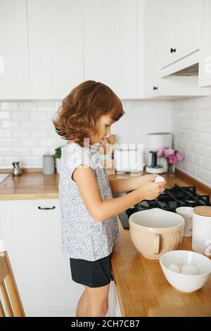 Bambina prepara un impasto per la cottura - rompe un uovo in una ciotola - cucina casalinga. Concetto di preparazione alimentare, cucina bianca su sfondo. Vita casual Foto Stock