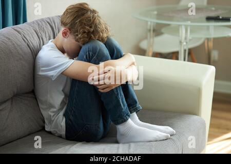 giovane ragazzo caucasico seduto da solo sul divano, faccia giù piangendo, a casa. ha problemi psicologici. bullismo cibernetico, problemi teen, solitudine Foto Stock