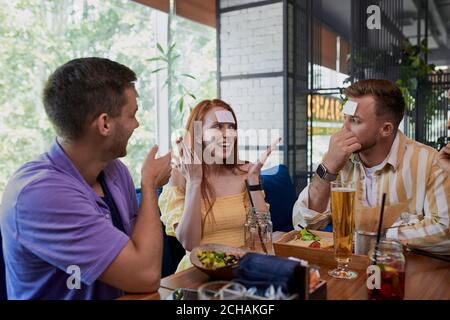 piccolo adesivo sulla fronte. amici che si divertono nel ristorante, caucasici giovani che giocano un hedbanz gioco, ridere Foto Stock