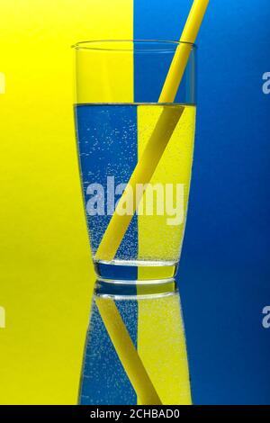 Un bicchiere d'acqua con una paglia che riflette uno sfondo blu e giallo. Foto Stock