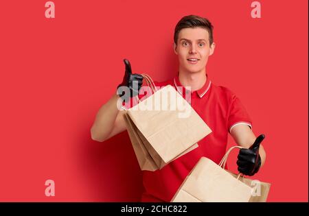 uomo in uniforme rossa con ordine di fast food isolato su sfondo rosso, corriere maschio contenente pacchetto di carta con cibo per i clienti, indossare guanti di protezione Foto Stock