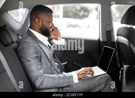 Giovane uomo d'affari nero che usa il telefono cellulare e il computer portatile in auto Foto Stock