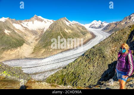 Donna escursionista con maschera chirurgica nella catena montuosa svizzera delle Alpi Bernesi in Vallese, Europa durante Covid-19. Ghiacciaio di Aletsch dal punto panoramico di Eggishorn Foto Stock