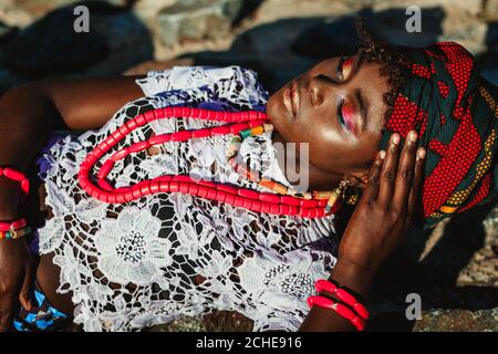 Foto di moda di giovane donna africana modello in costume nazionale, Turban e colorful brillante compongono sdraiato sulle pietre. Concetto turistico esotico Foto Stock