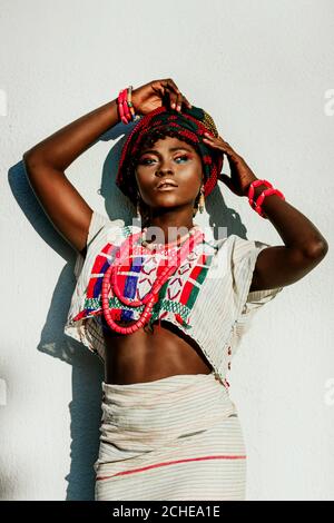 immagine ritagliata del modello femminile africano di moda in costume nazionale, gioielli, turbante e compongono in posa su sfondo bianco parete, sole giorno. Foto Stock