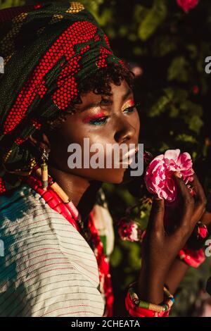 immagine ritagliata del modello femminile africano di moda in costume nazionale, gioielli, turbante e compongono in posa in giardino di rose, luminoso sole giorno. Foto Stock