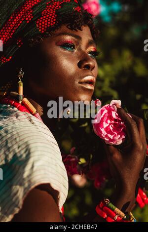 immagine ritagliata del modello femminile africano di moda in costume nazionale, gioielli, turbante e compongono in posa in giardino di rose, luminoso sole giorno. Foto Stock