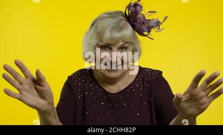 Ritratto di una donna bionda anziana e amichevole che agita le mani guardando la macchina fotografica con un sorriso toothy, dicendo Ciao, saluto. Anziana elegante nonna donna su sfondo giallo in studio Foto Stock
