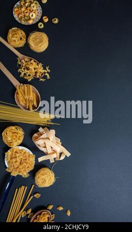 Concetto di cucina della pasta. Pasta cruda varie forme e cucchiai su fondo in pietra nera, vista dall'alto Foto Stock