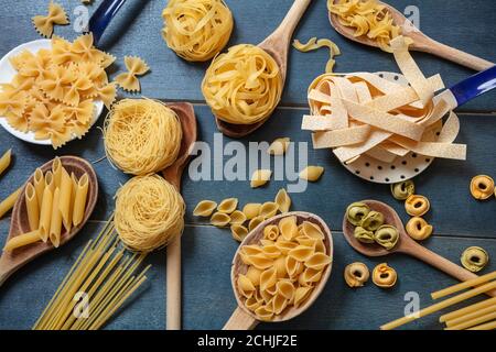 Concetto di cucina della pasta. Pasta cruda varie forme e cucchiai piatti posati su sfondo di tavola di legno blu, vista dall'alto Foto Stock