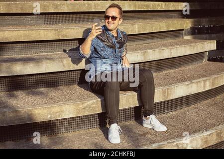 Sorridere uomo in occhiali da sole sta scattando foto utilizzando lo smartphone mentre si è seduti sulle scale. Giovane caucasico maschio circa 27 anni fa le immagini. Foto Stock