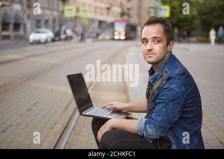 Un giovane uomo pensivo con un taccuino in mano è seduto vicino alla pista ferroviaria sullo sfondo di un tram a cavallo. Foto Stock