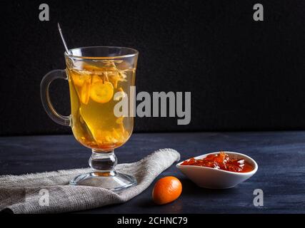 Tè al gelsomino con kumquat in una tazza di vetro su una tavola di legno su sfondo nero. Primo piano, copia spazio. Foto Stock