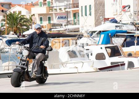 Vodice, Croazia - 1 settembre 2020: Uomo anziano che guida uno scooter elettrico a ruota larga con un sedile, sul molo di una piccola città mediterranea Foto Stock