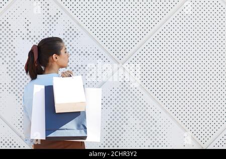 Vista posteriore ritratto di giovane donna che tiene borse per la spesa mentre si sta in piedi su sfondo bianco testurizzato, spazio di copia Foto Stock