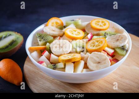 Insalata vegetariana di banane, mele, pere, kumquat e kiwi su sfondo boschivo nero, primo piano, fuoco selettivo. Foto Stock