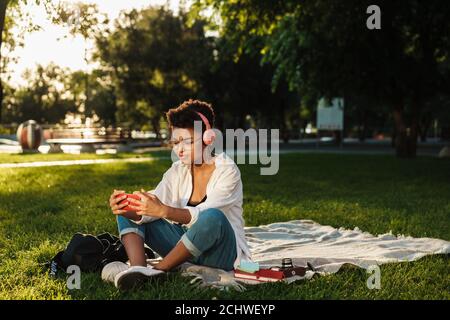 Immagine di una donna africana concentrata seduta all'aperto nel parco un'erba mentre si ascolta la musica con le cuffie e si usa il cellulare telefono Foto Stock