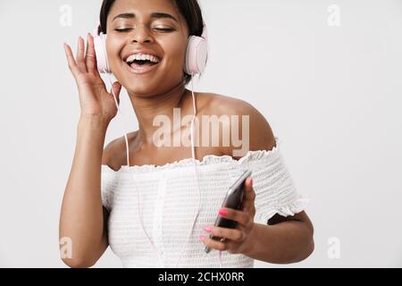 Immagine di gioiosa donna afroamericana in cuffia con cellulare mentre cantava isolato su parete bianca Foto Stock