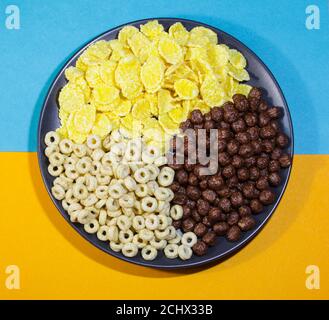 Anelli di miele, cornflakes e palle di cioccolato in un piatto nero su uno sfondo colorato. Concetto di cibo sano. Vista dall'alto. Foto Stock