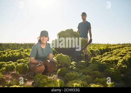 Ritratto a lunghezza intera di due sorridenti lavoratori che guardano la macchina fotografica mentre posano con la raccolta a piantagione di verdure all'aperto, copia spazio Foto Stock