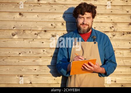 Vita in su ritratto di uomo bearded maturo che tiene gli appunti e che indossa il grembiule mentre in piedi contro sfondo rustico di legno alla luce del sole, spazio di copia Foto Stock