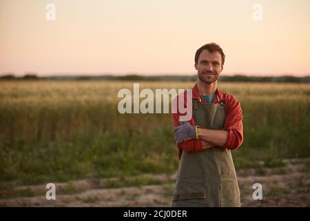 Vita in su ritratto di giovane agricoltore che posa con sicurezza con le braccia incrociate mentre si trova in campo al tramonto e sorridendo alla macchina fotografica, copia spazio Foto Stock