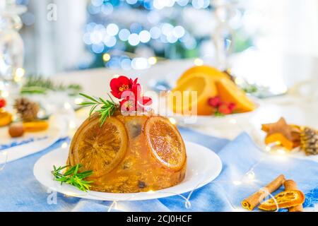 Tradizionale budino di Natale inglese decorato con arance e fiori in un ambiente festoso tavolo con frutta, spezie e luci di albero di Natale Foto Stock