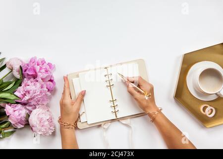 Le mani di donna ben curate sta scrivendo nel diario con i rifornimenti di cancelleria set onside e bouquet di peonie Foto Stock