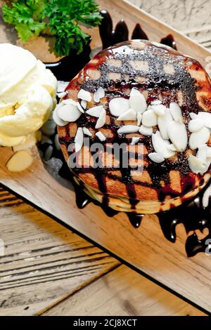 Vista dall'alto dei pancake alla banana con noci di macadamia e sciroppo di cioccolato sulla pila di frittelle e gelato alla vaniglia su un piatto di legno su un tavolo di legno. Foto Stock