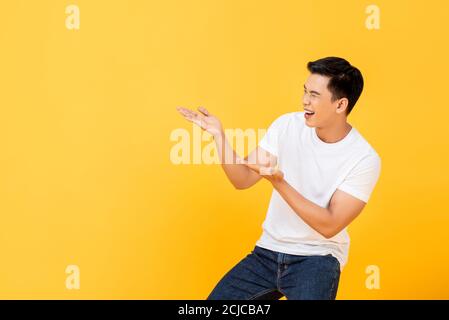 Divertente Ritratto di sorridere giovane bell'uomo asiatico presentandosi con apri il gesto delle palme in uno sfondo giallo isolato dello studio con copia spazio Foto Stock