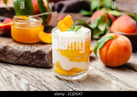 Dessert alla frutta di pesca in una tazza di vetro su un tavolo rustico in legno con frutta fresca di pesca, marmellata di pesche. Dessert fatto in casa con frutta. Macedonia di frutta con yogurt o. Foto Stock