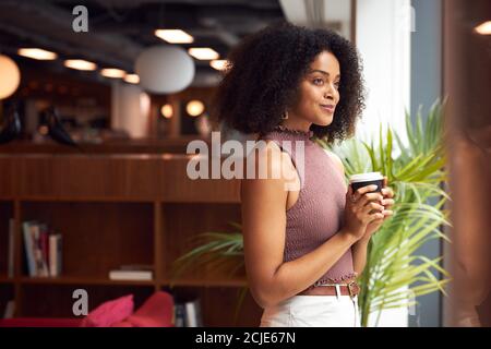 Giovane Businesswoman da finestra con caffè Takeaway prendere UNA pausa Nel moderno ufficio open space Foto Stock