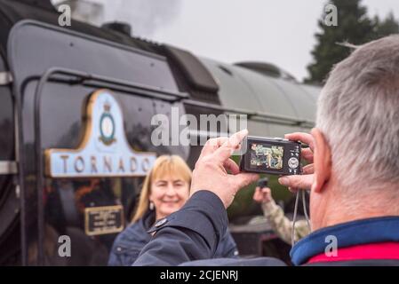 Uomo di mezza età che fotografa sua moglie, signora in piedi di fronte alla locomotiva a vapore britannica Tornado 60163. Foto Stock