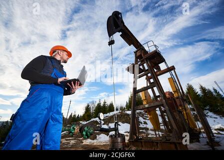 Basso angolo di operatore del petrolio che tiene il notebook e guarda il martinetto della pompa del petrolio. Operatore di petrolio in tute da lavoro, casco in piedi vicino all'unità di pompaggio dell'olio. Concetto di industria petrolifera, estrazione dell'olio. Foto Stock