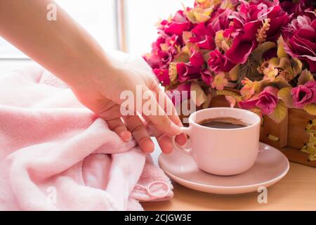 La mano di una donna raggiunge la sua tazza rosa di caffè nero vicino al fiore rosa sul legno texture background.International Coffee Day. Foto Stock