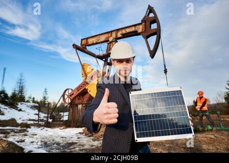 Uomo d'affari che tiene un pannello solare portatile, mostrando un gesto di approvazione. Tecnico che lavora con il martinetto della pompa sul territorio del campo dell'olio in background. Concetto di industria petrolifera, fonti energetiche alternative. Foto Stock